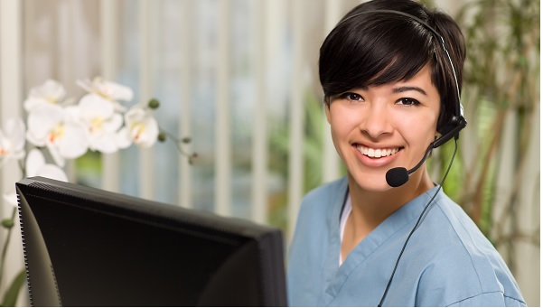 woman in scrubs at computer smiling and wearing headset